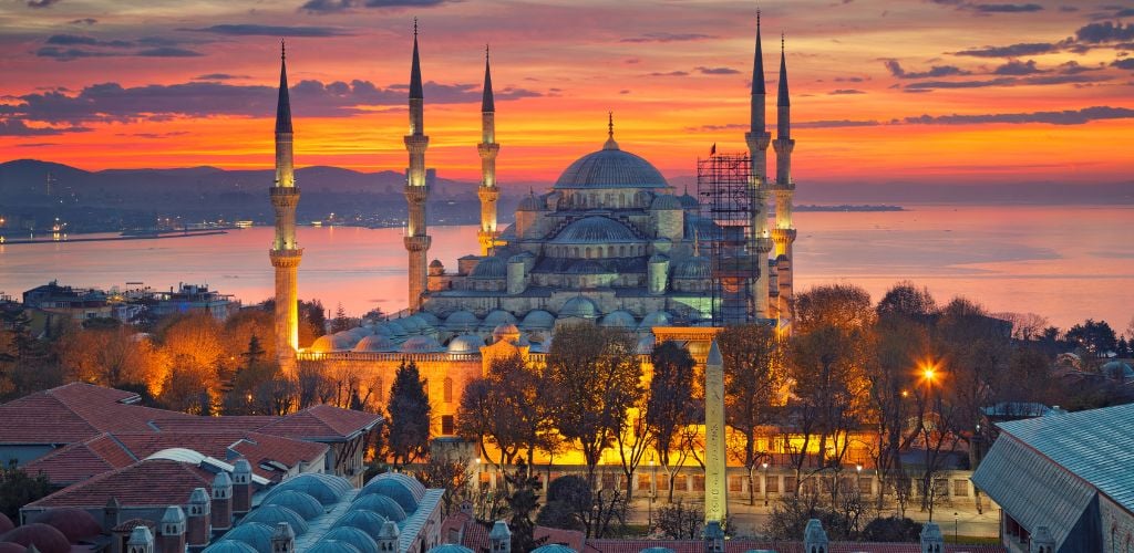 On a sunset atmosphere, an old mosque, surrounded by smaller building structures and a few trees.